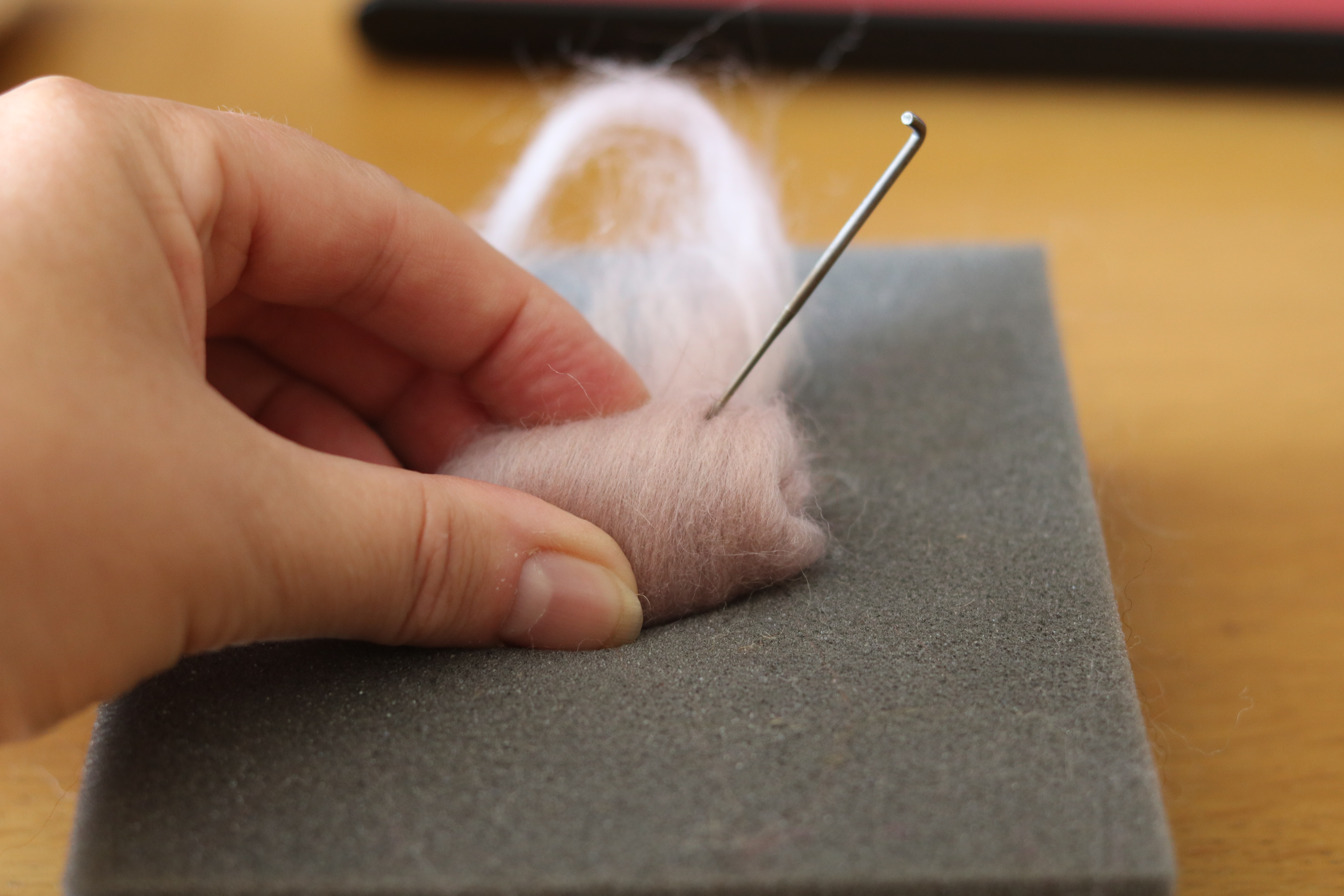A close-up of a hand felting a small, pale pink wool shape with a needle on a grey foam block, with wool fluff scattered around and a blurred background.