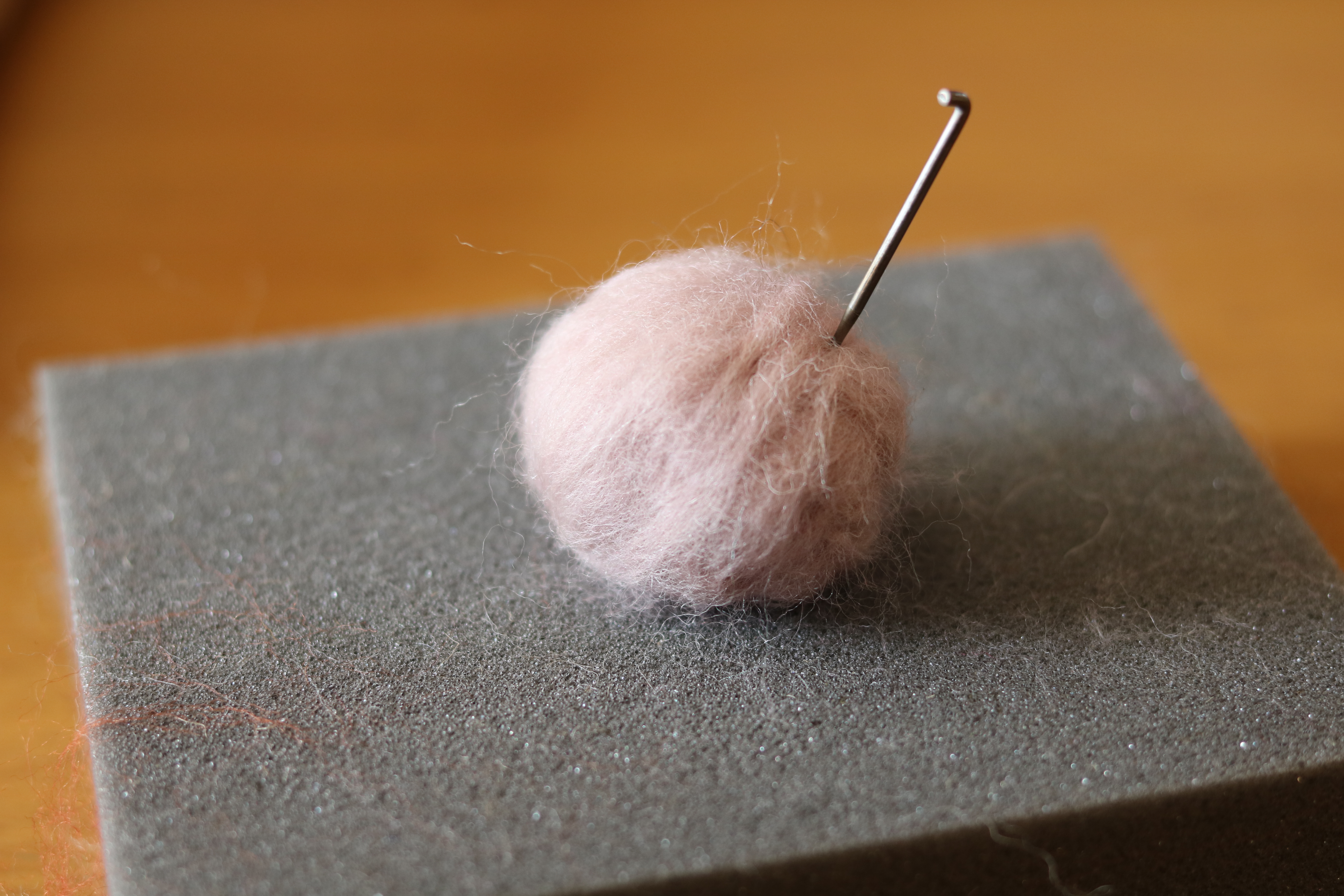 A pink wool needle felted ball with a metal felting needle inserted, placed on a gray foam block. the surface shows traces of wool fibers.