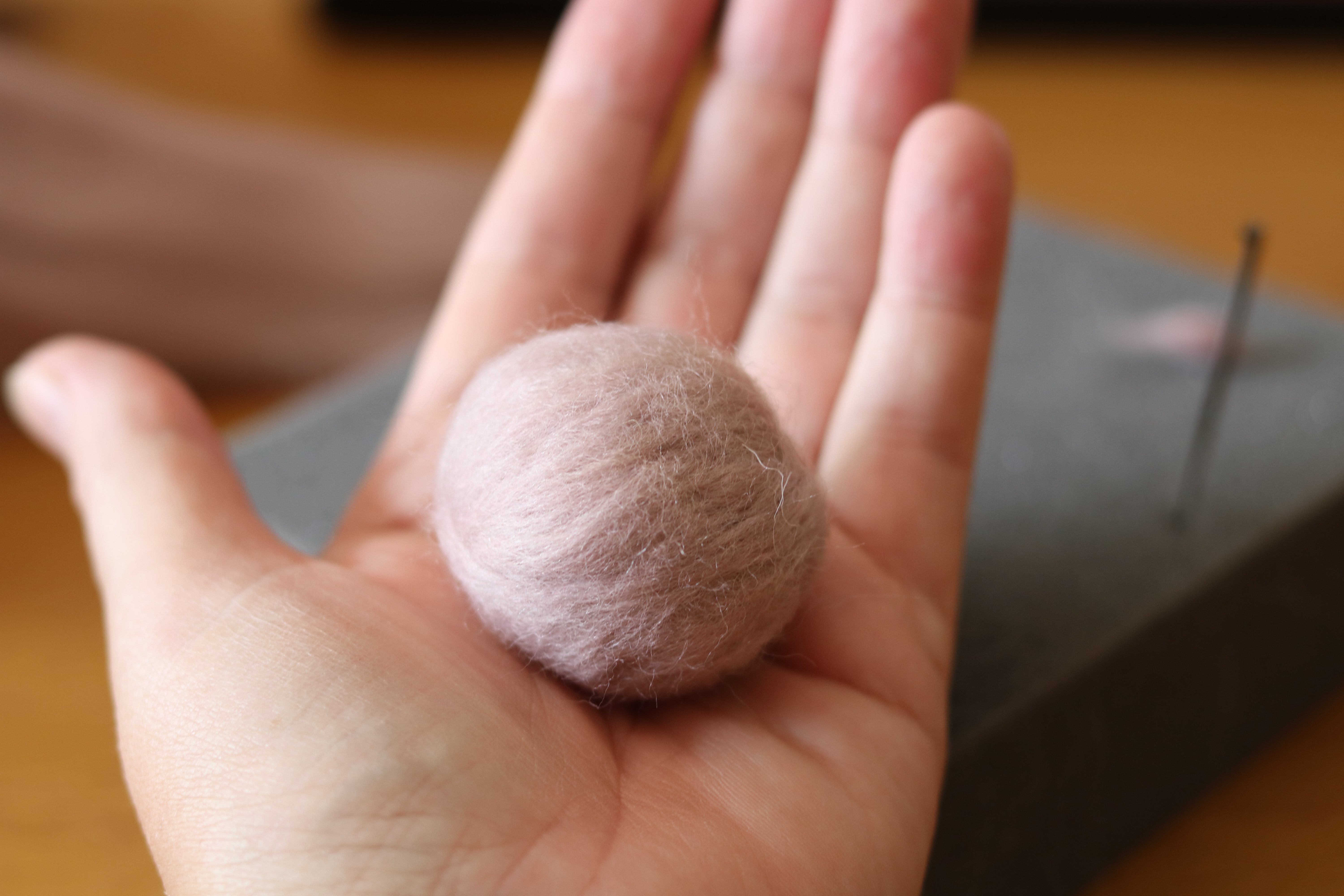 A person's hand displaying a small, round, pink felted wool ball, with a needle felting pad and felting needle in the background on a wooden surface.