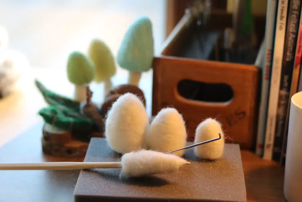 A crafting table with materials for needle felting, featuring partially made white and brown wool shapes, forests and a small box labeled "pines." natural light illuminates the scene.