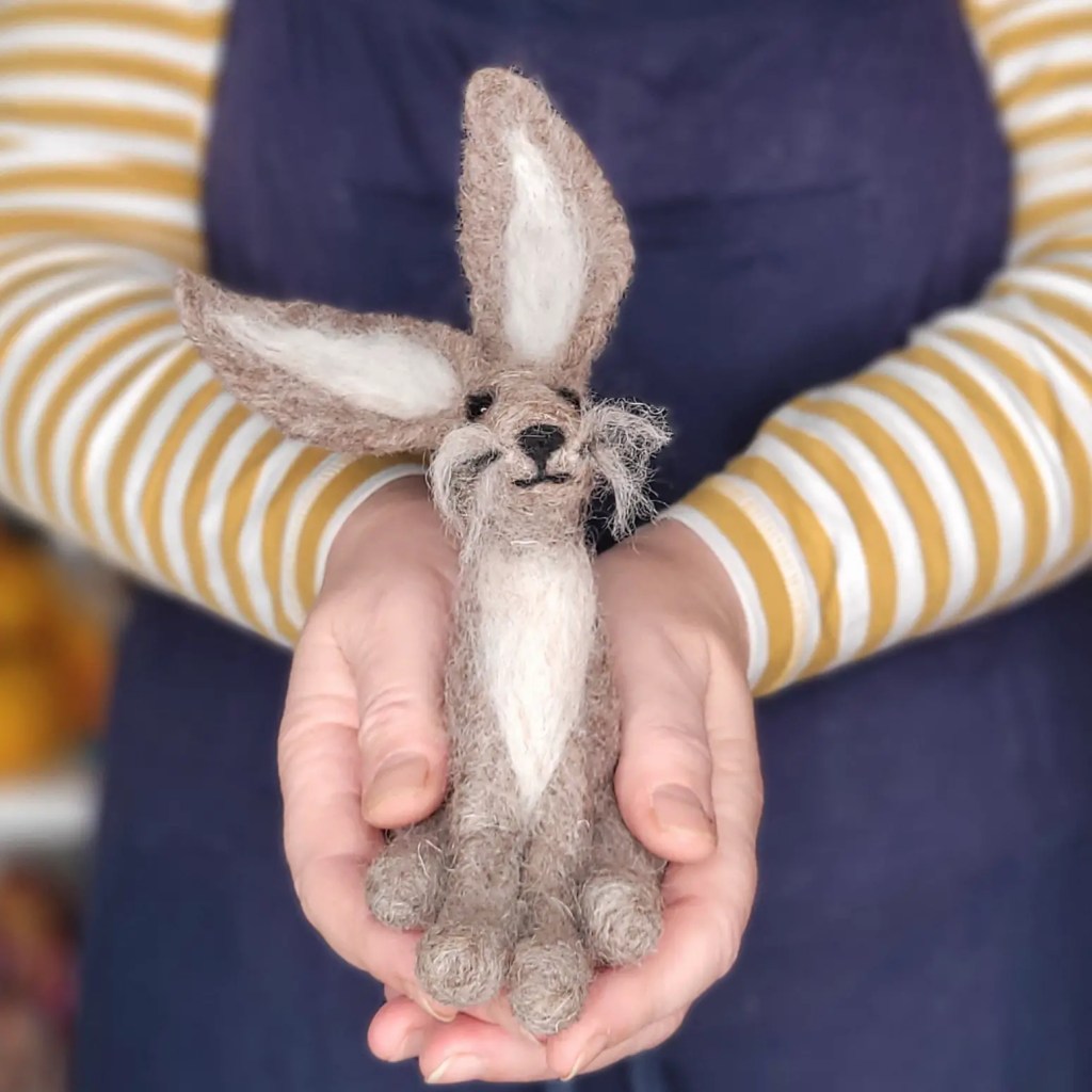 A person wearing a striped yellow and blue shirt holds a small, needle felted bunny with floppy ears and a whimsical expression, presented against a soft-focus background.