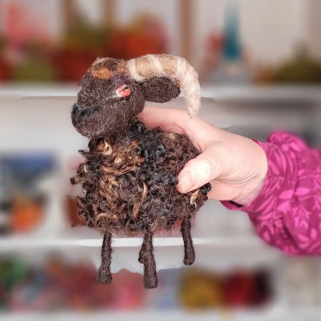 A hand holds a small, intricately crafted needle felting sculpture of a sheep with a dark body, curly fleece, and distinct beige horns, set against a blurry background of colorful shelves.