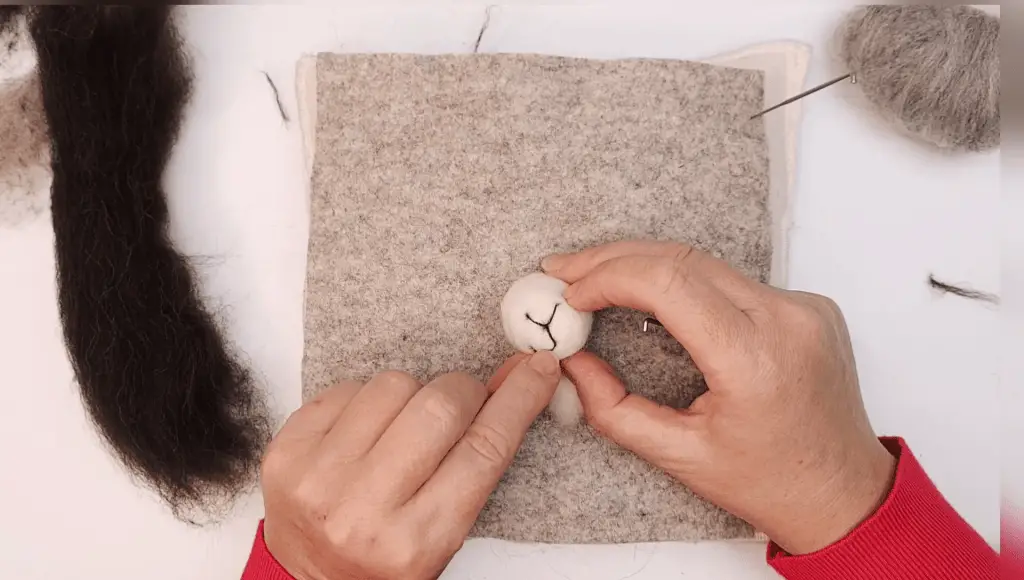 Hands crafting a small white felt object on a grey felt mat with wool and felting tools nearby. the object being made has a simple face drawn on it.
