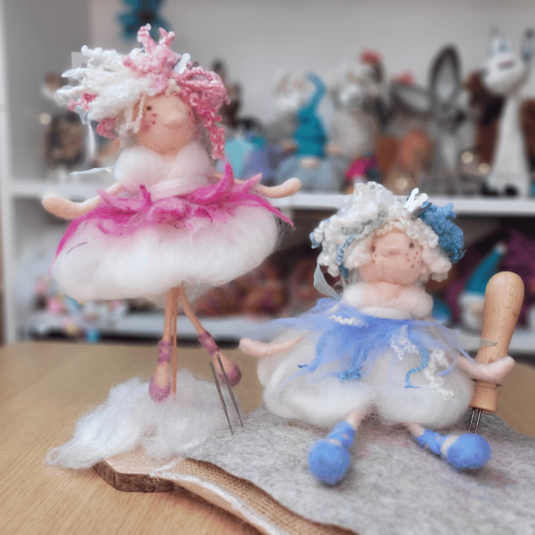 Two handcrafted felt dolls with colorful curly hair and fluffy dresses, one in pink standing on a wooden base and the other in blue sitting on felt with a felting needle. They are in a cozy, craft-filled room.