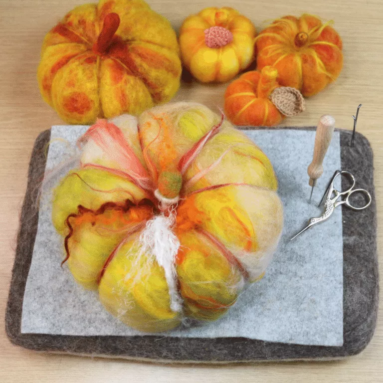 Colorful felted wool pumpkins, felting tools, and scissors on a foam mat atop a wooden surface; one pumpkin is being shaped with felting needles.